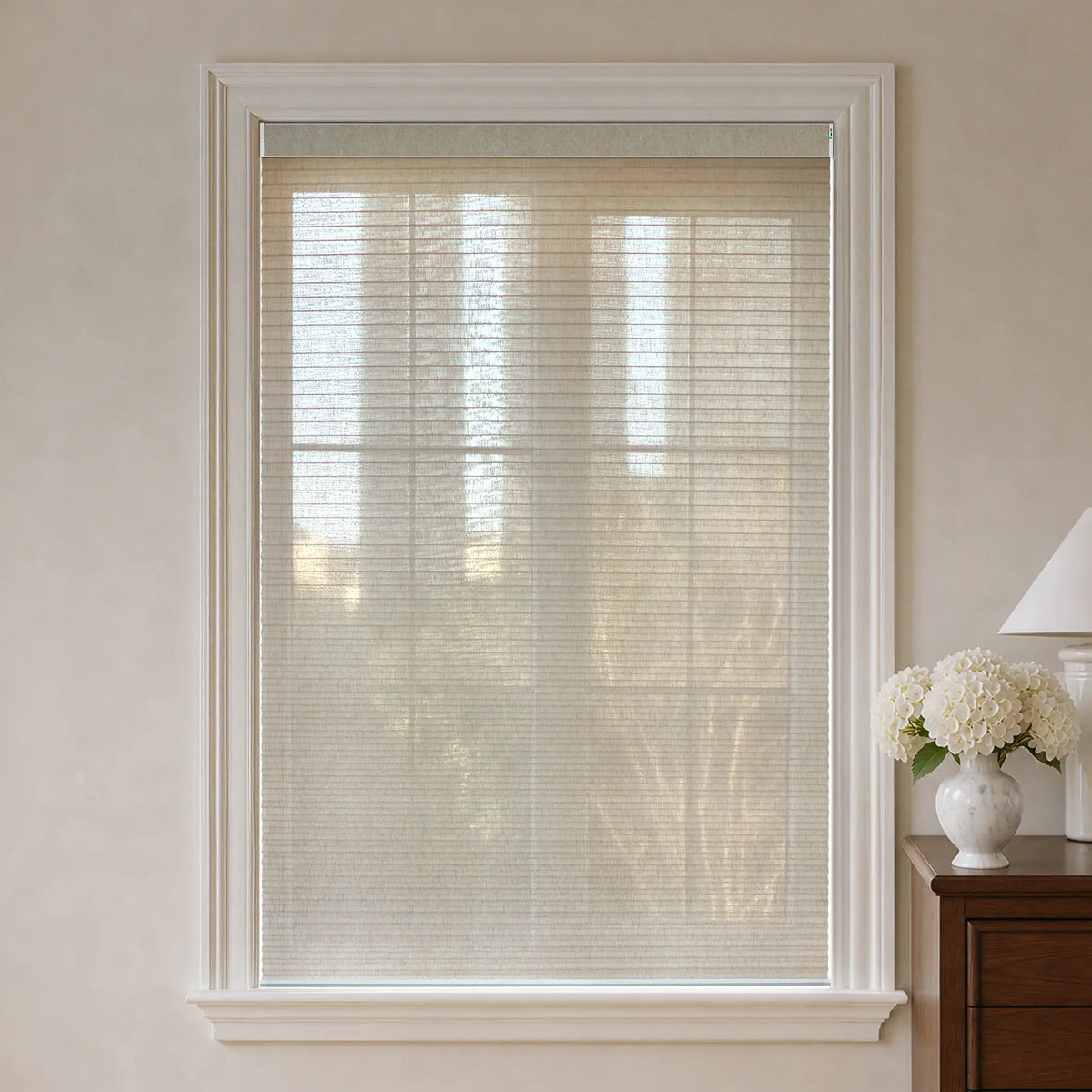 Beige motorized cellular shades with a white frame installed on a window in a neutral-toned room.