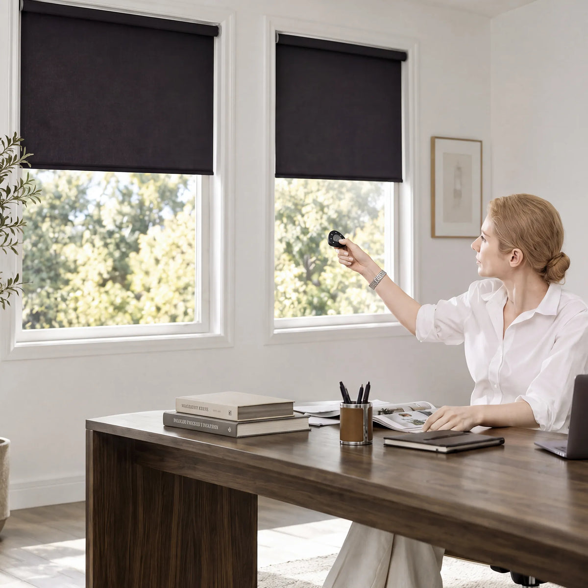 A woman in a white blouse sits at a wooden desk in a bright home office, reaching toward two black motorized no-drill roller shades with a remote. The room has white walls, large windows with green foliage outside, and books and office items on the desk.
