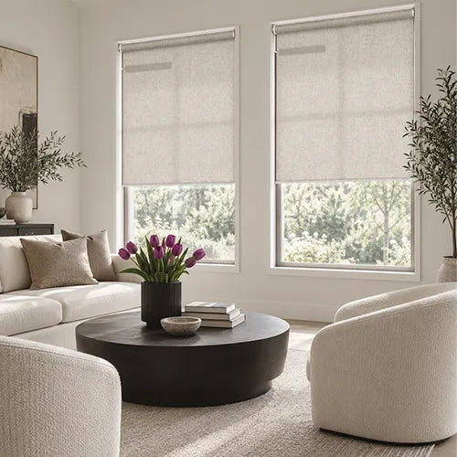 Two beige motorized no-drill roller shades  windows in a bright, modern living room with a white sofa, textured armchairs, and a round black coffee table.