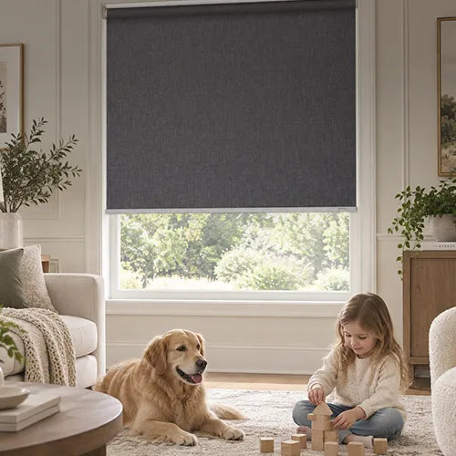 A dark charcoal gray motorized no-drill roller shades with a textured fabric cover, shown partially drawn on a window in a bright, modern living room. A golden retriever dog is lying on the rug next to a young girl who is sitting and playing with wooden blocks.