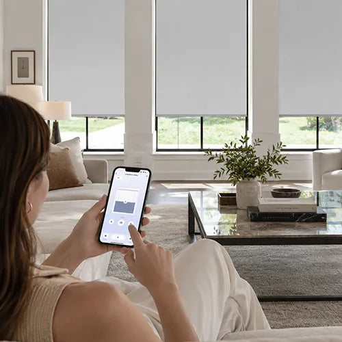 A woman sits on a white sofa in a bright living room, holding a smartphone that displays a smart home app controlling the motorized no-drill roller shades  on large windows behind her.