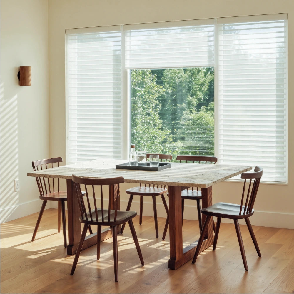 A dining room scene with a rectangular wooden table and four matching wooden chairs in front of large windows with white horizontal sheer blinds and a view of green trees outside.