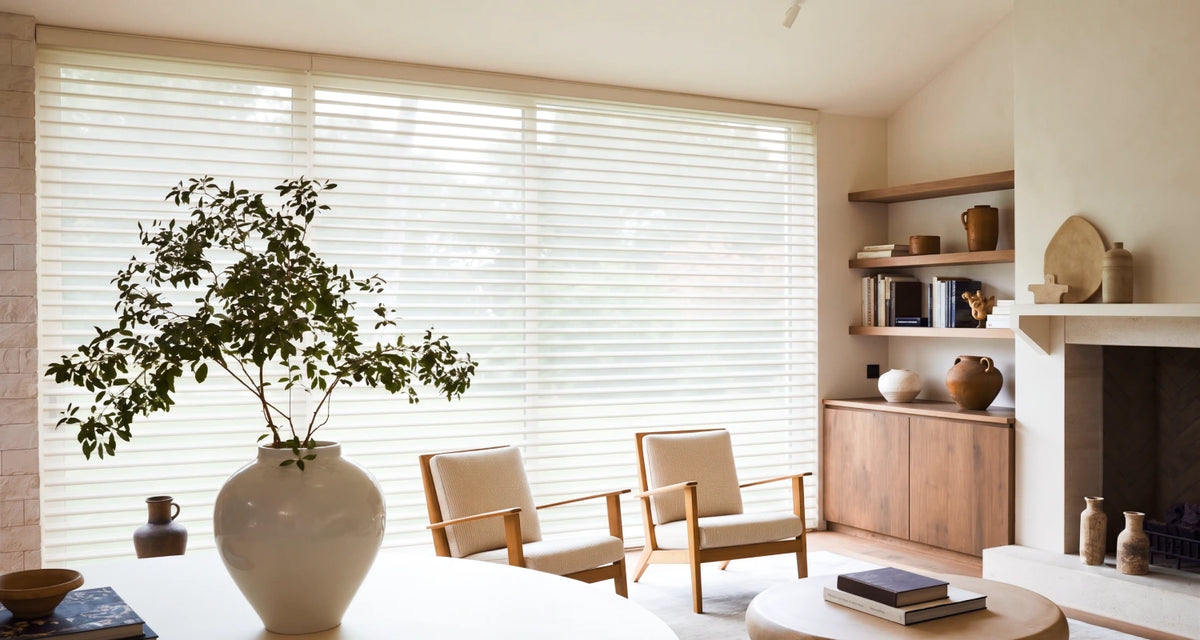 A bright, modern living room with large cream horizontal sheer blinds covering a window, styled with two wooden armchairs, a white ceramic vase with a leafy plant, and a fireplace with wooden shelves.
