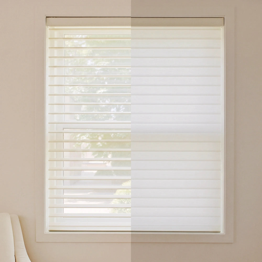 White shangri-la sheer blind shades partially drawn over a window, shown in a split view with and without light filtering through. The window is set in a beige wall, and a small portion of a chair is visible in the bottom left corner.