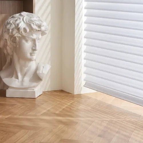A white classical-style bust sculpture resembling Michelangelo's David, displayed on a light wood herringbone floor in a sunlit room with white shangri-la sheer blinds.