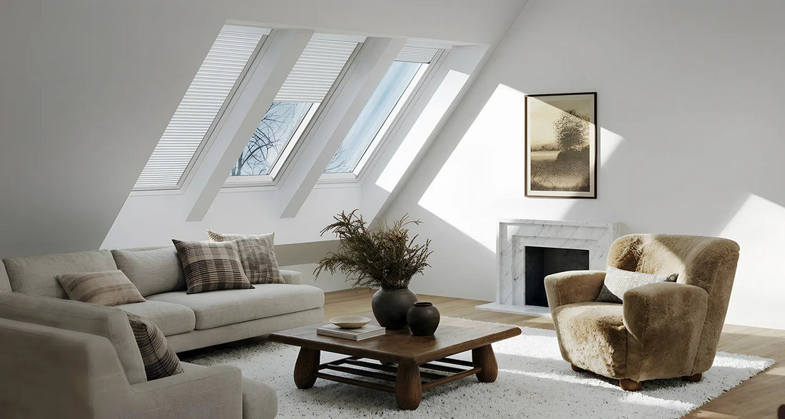 A bright, modern attic living room with a light beige sofa, a brown wooden coffee table, a plush beige armchair, and a white fireplace. Two large skylights with white blinds let in natural sunlight, casting soft shadows on the walls.