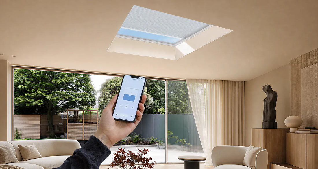 A hand holds a smartphone showing a smart home app interface, controlling a rectangular skylight installed on the ceiling of a modern beige living room with large windows and outdoor greenery visible.