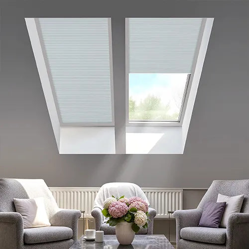 Two white cellular sun and shade curtains installed in a slanted attic window of a gray living room.