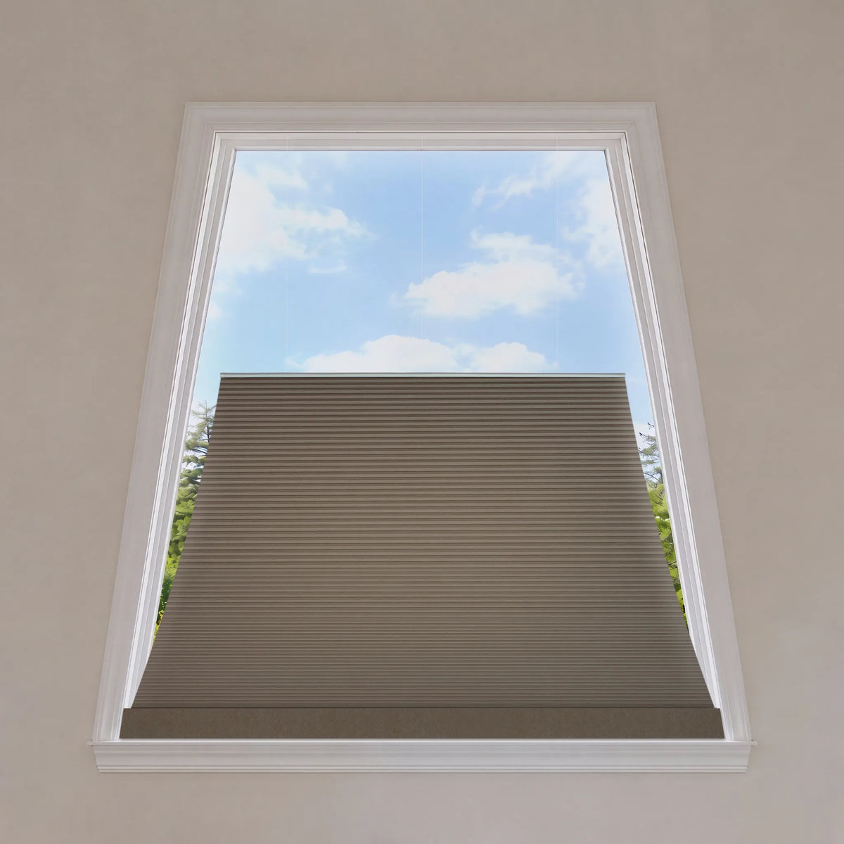 A beige trapezoid window shade partially drawn down inside a white-framed window, with a view of blue sky and green trees outside.