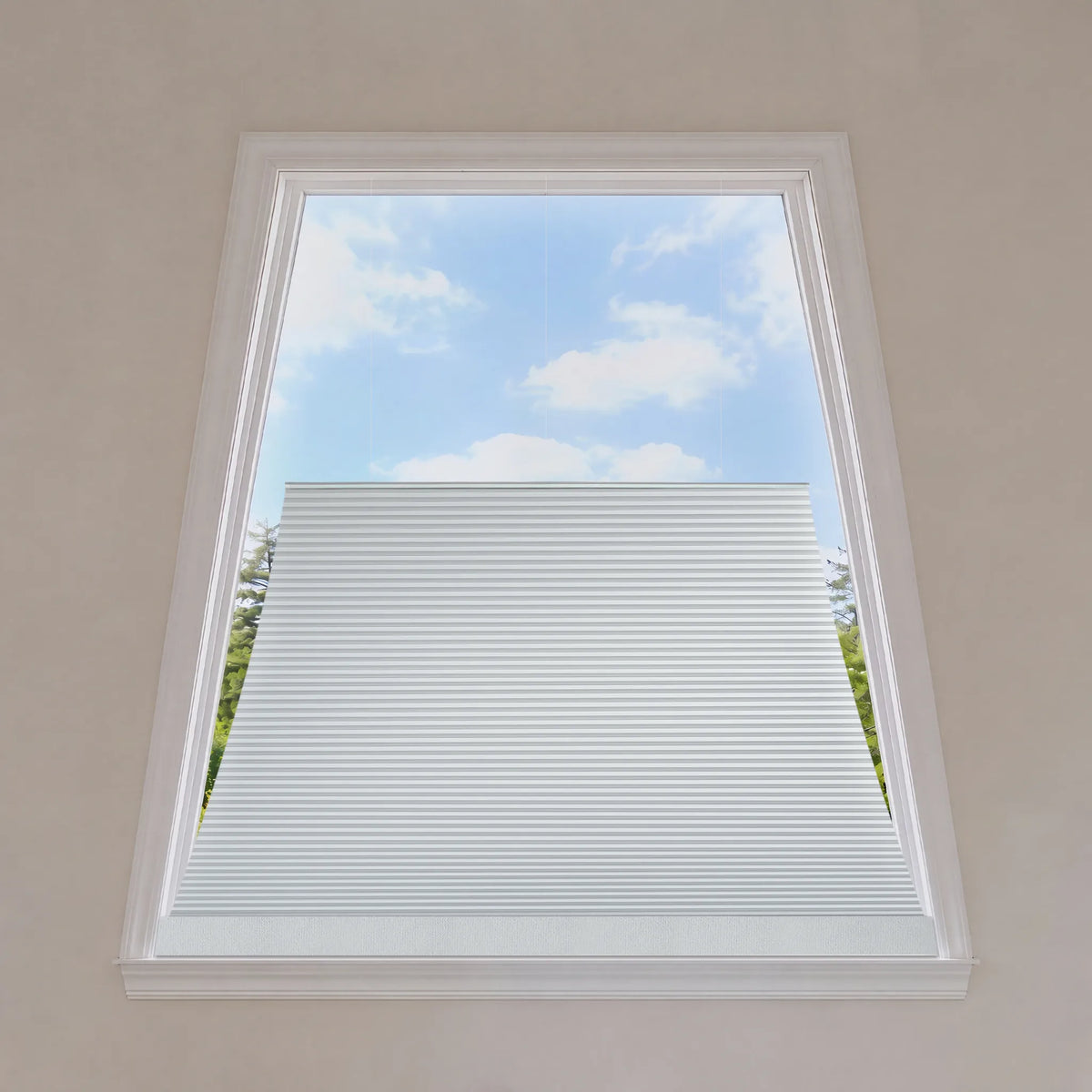 White cellular window shade installed in a tall rectangular window, shown from inside the room looking out toward a blue sky with clouds.