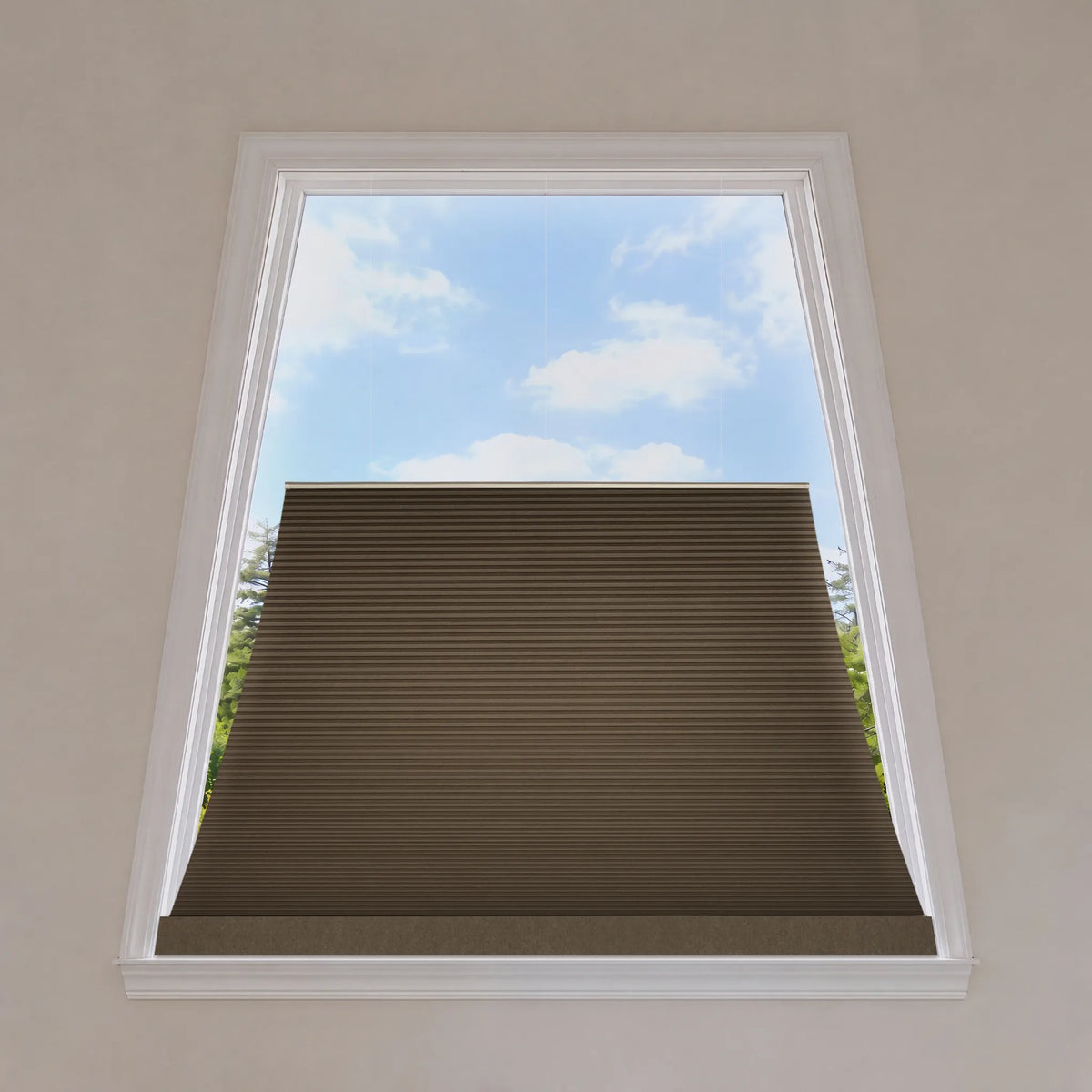 A brown trapezoid window shade partially drawn down inside a white-framed window, with a blue sky and green trees visible outside.