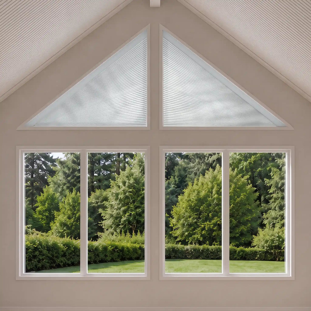 A set of white triangular cellular window shades installed in a gabled attic window, shown from a top-down perspective with a lush green garden visible outside.