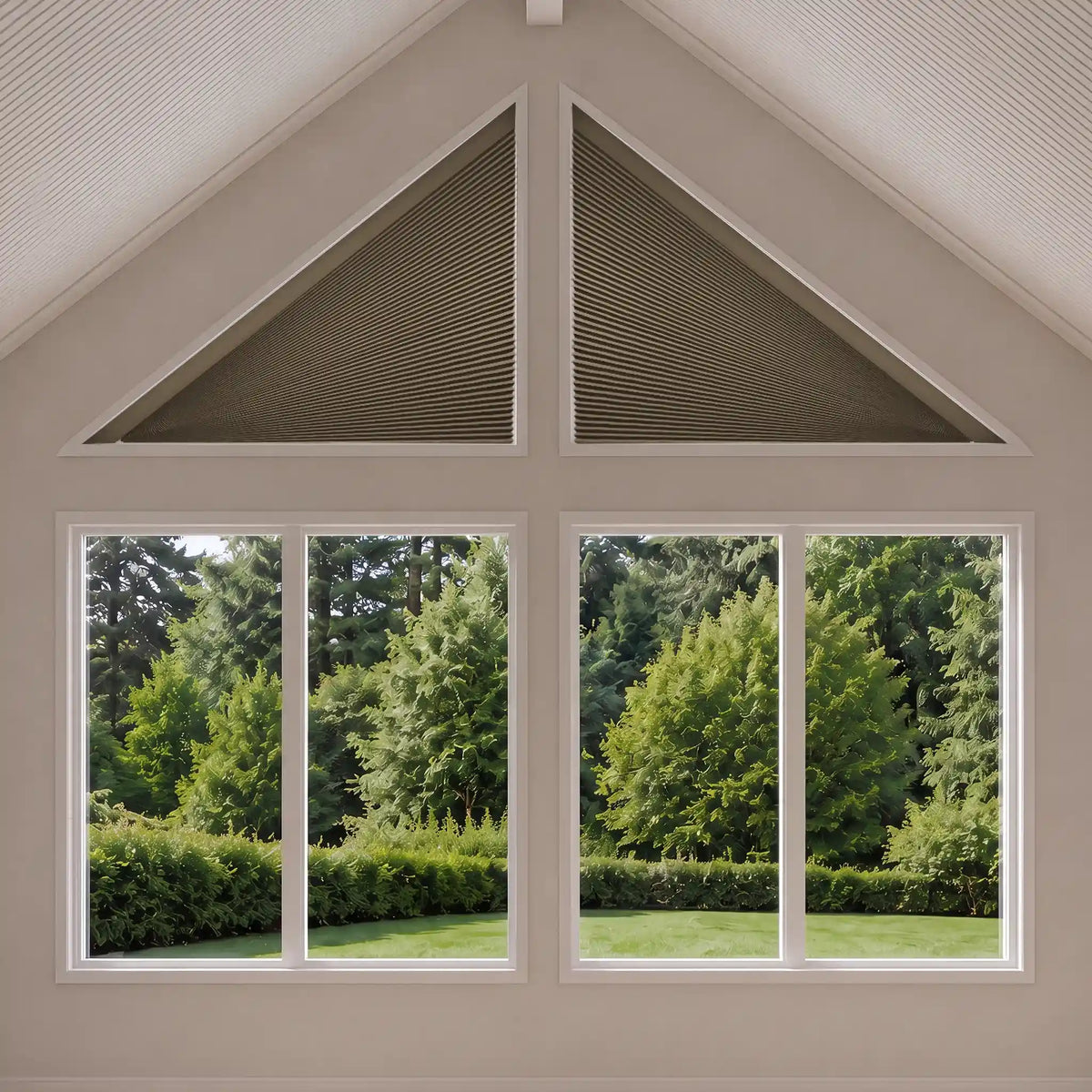 Triangular cellular shade installed in the gable above a multi-pane window, showing a green garden outside. The shades are a dark taupe color and the room has light beige walls.