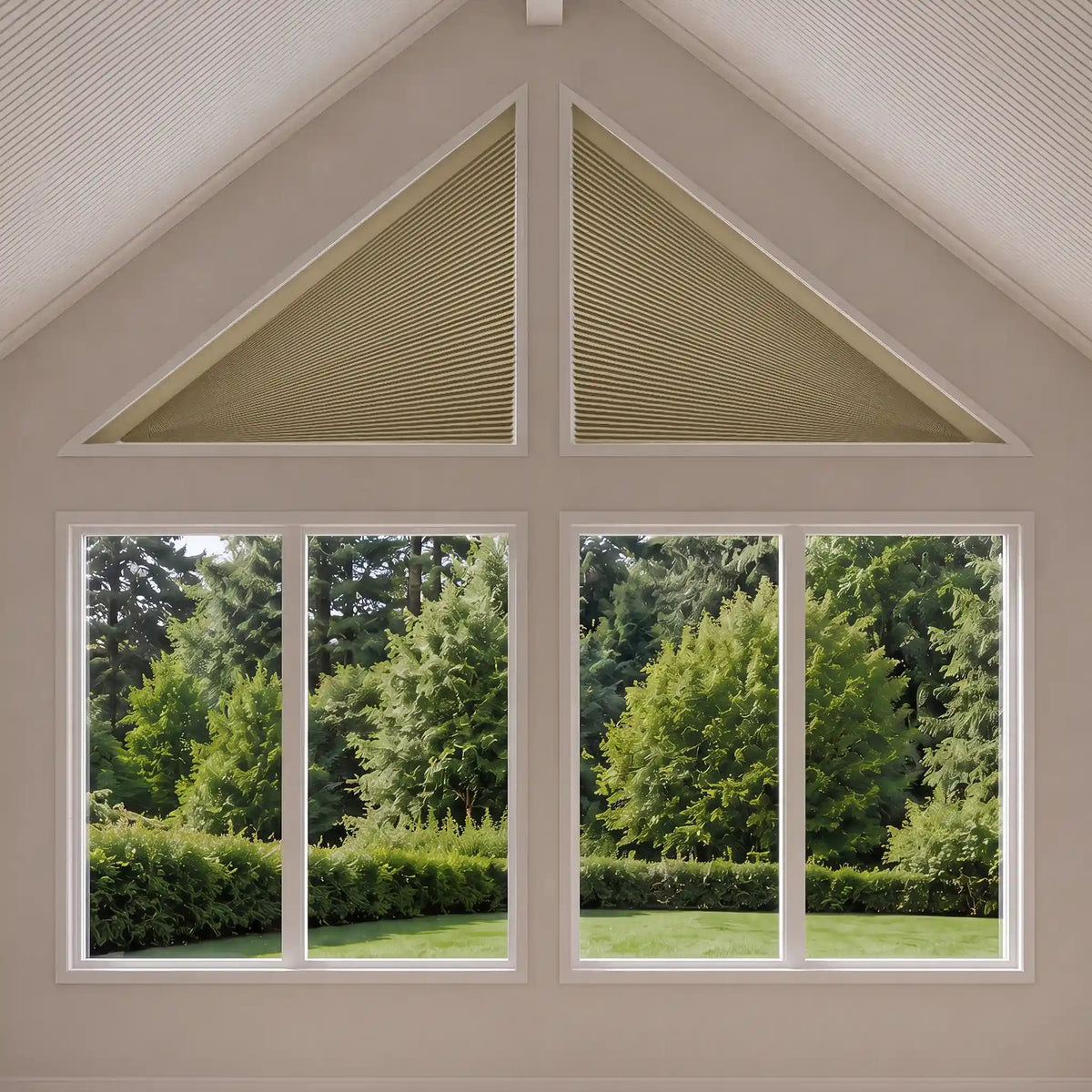 Triangular cellular shade window blinds in a light beige color installed on a gable wall above two large multi-pane windows showing a green garden view.