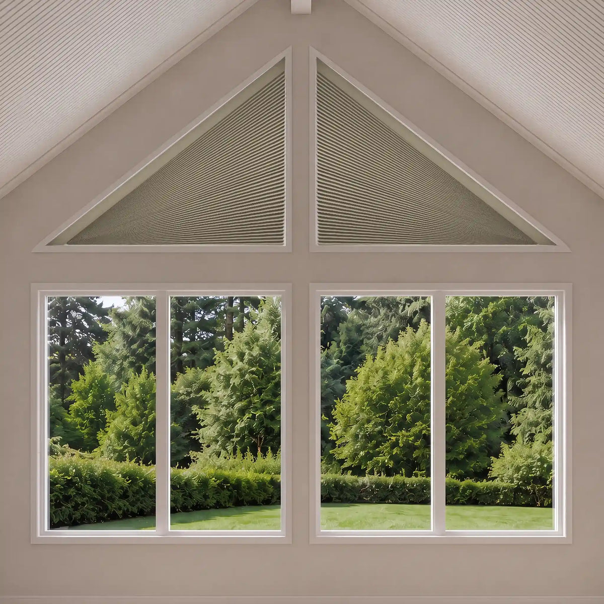 Triangular cellular shade window blinds in a light beige color installed on a gable wall above two large multi-pane windows showing a green garden view.