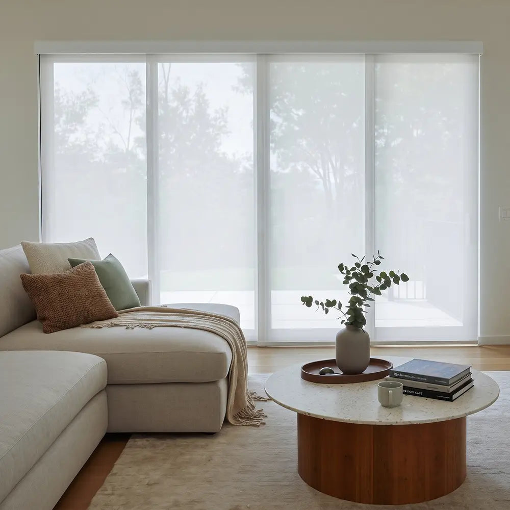 A modern living room with a beige sectional sofa, a round wooden coffee table, and large lignt filter  panel track blinds covering tall windows in the background.
