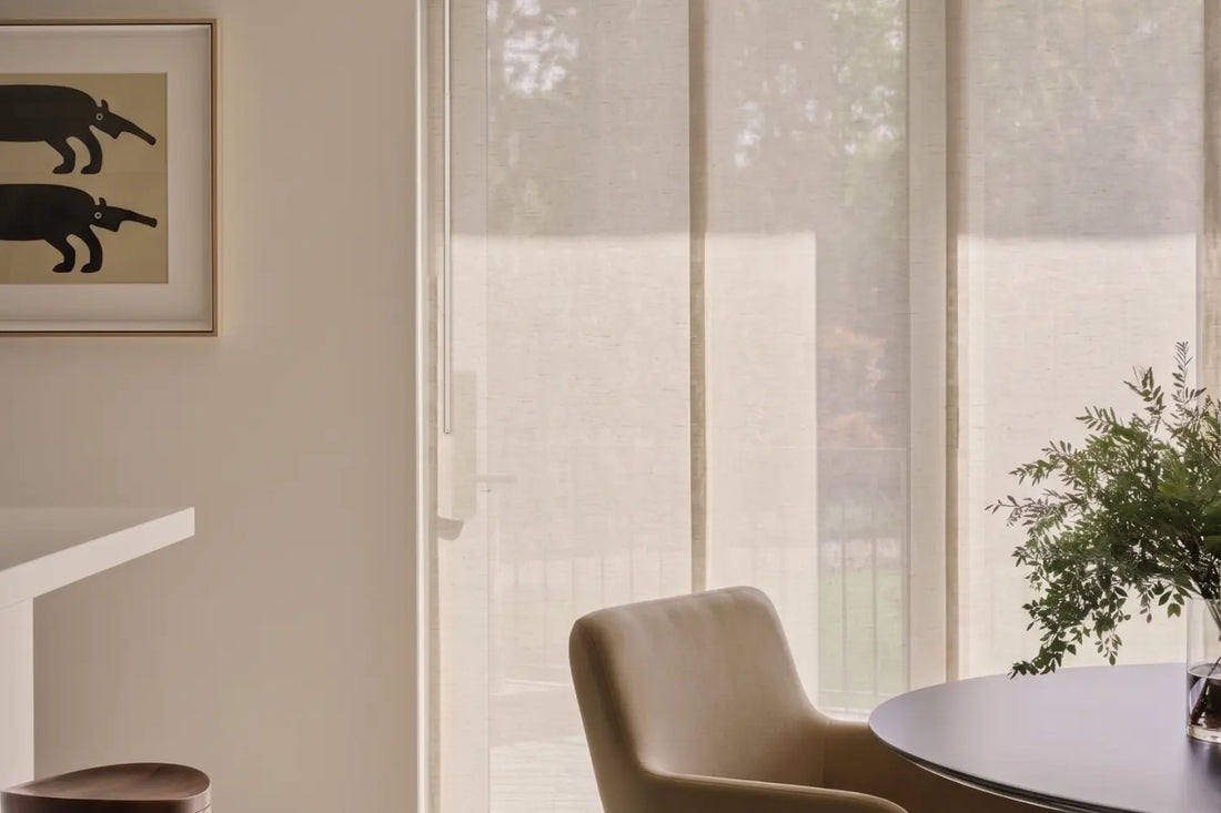 A modern interior scene with manual panel track blinds in a warm beige tone, shown installed on a window in a minimalist room with a chair, table, and framed wall art.