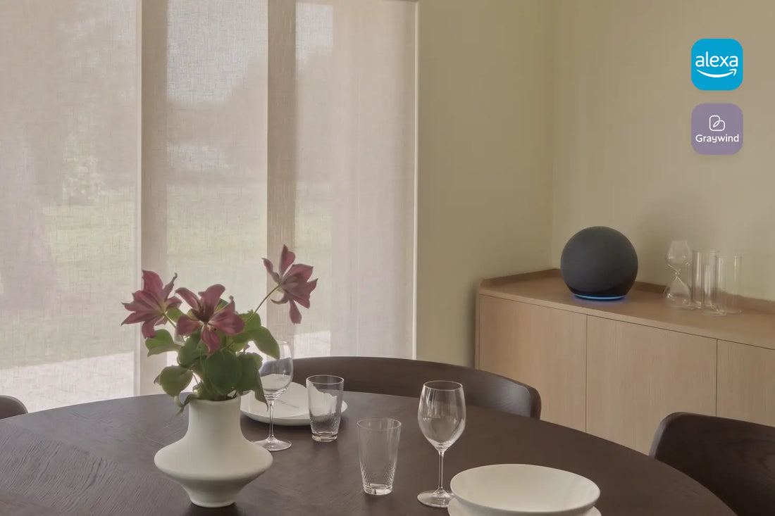 A modern dining room scene with a round dark table set with white plates, wine glasses, and a white vase holding pink flowers. A light wood sideboard stands against a beige wall with smart panel track blinds, and a alexa echo speaker sits on top.