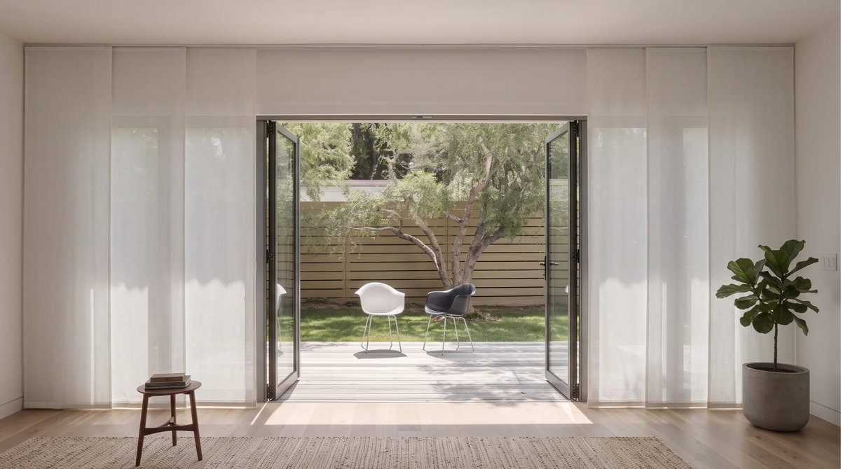 A modern interior room with floor-to-ceiling glass bi-fold doors opening onto a backyard patio with two chairs and a tree. The room has white textured panel track blinds, light wood flooring, a small wooden stool, and a potted plant.