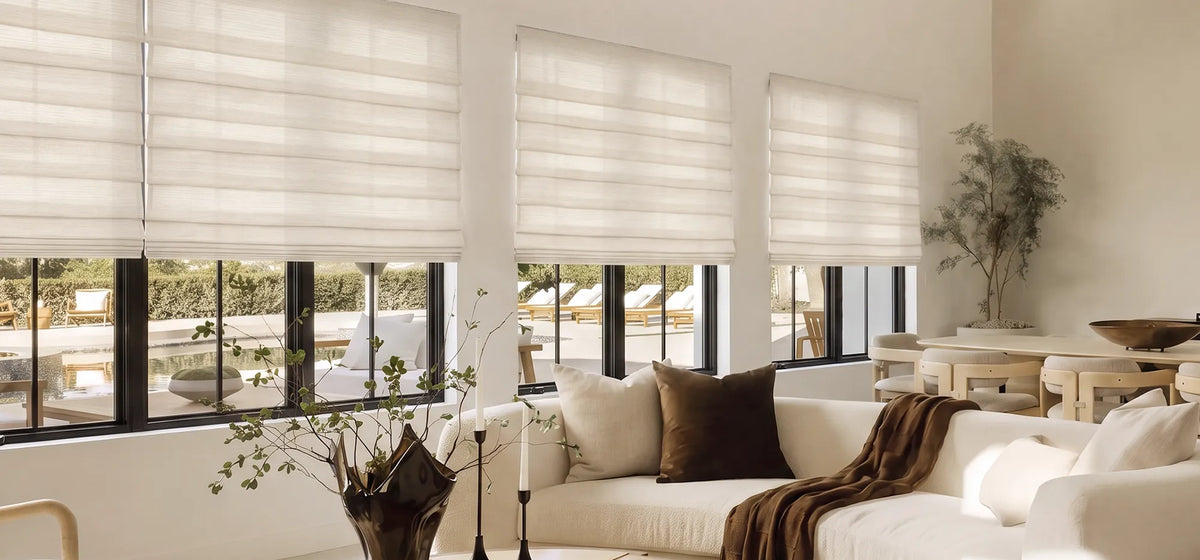 Light-colored soft fold Roman shades across wide living room windows above a sofa. 