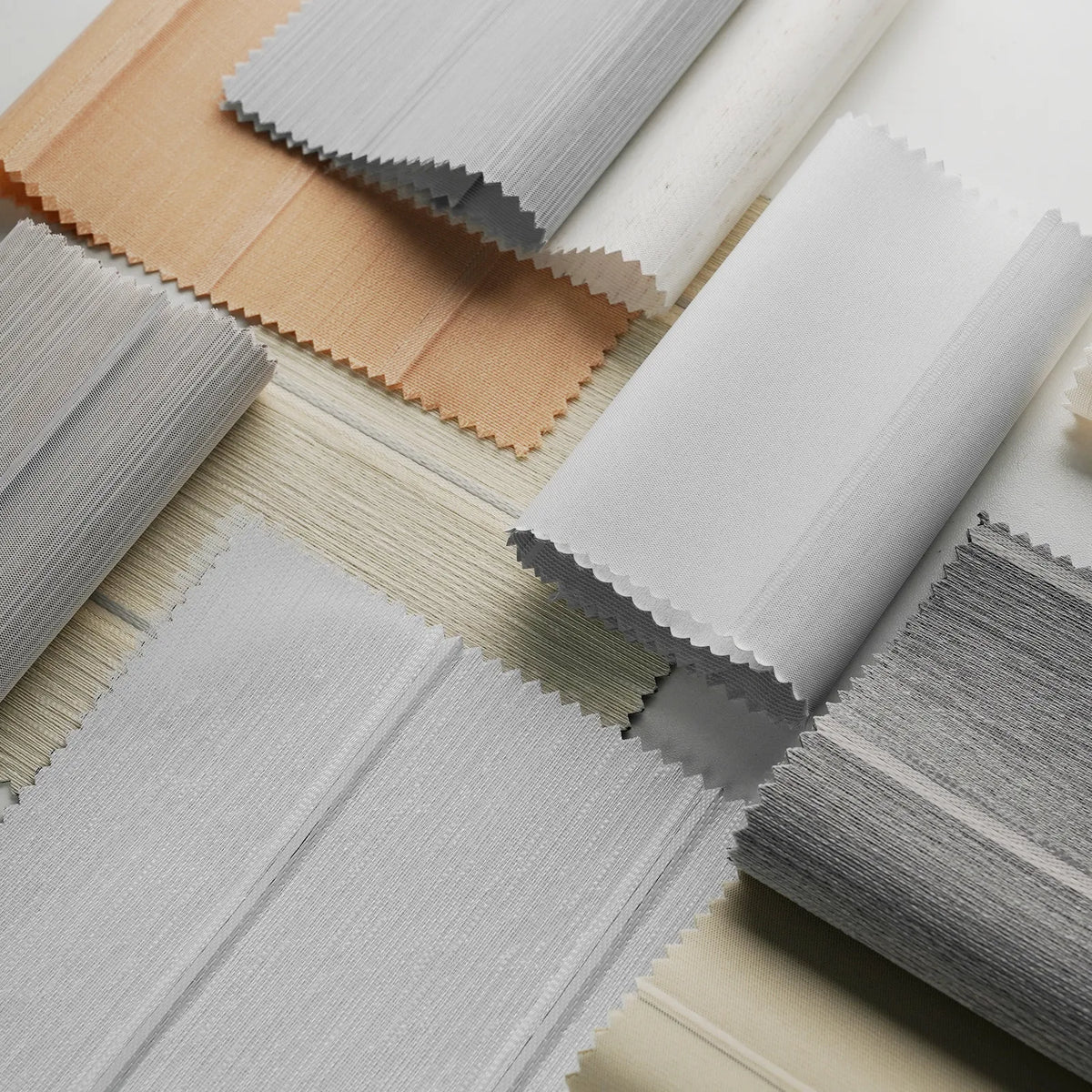 A close-up arrangement of layered fabric or material swatches in white, light gray, beige, and taupe, each folded with scalloped edges, displayed on a light surface.