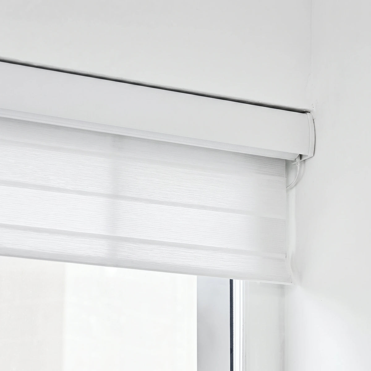 A close-up of a white shangrila shade partially drawn across a window in a bright, minimalist room.