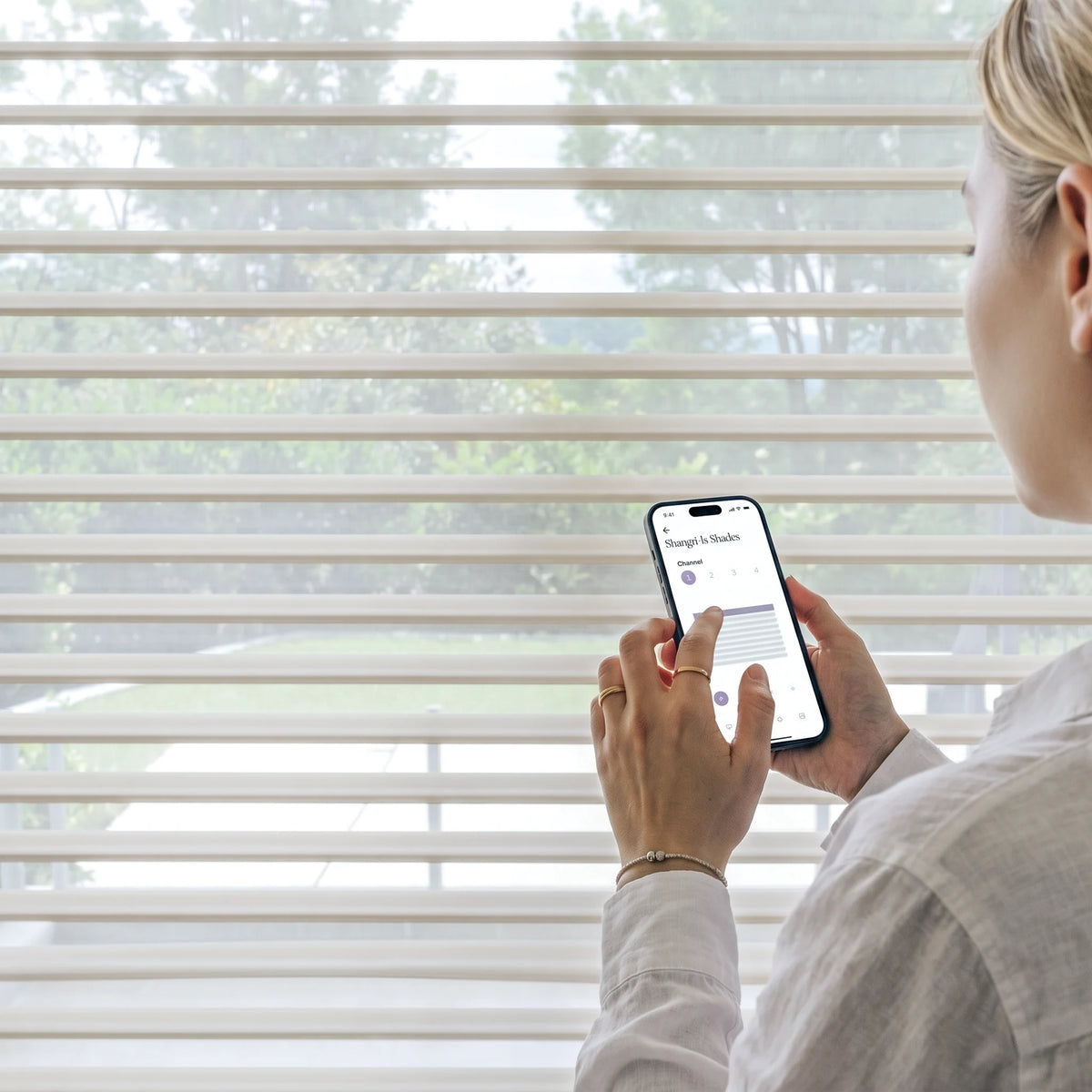 A woman in a light-colored shirt stands in front of window blinds, holding a smartphone that displays a smart home app interface.