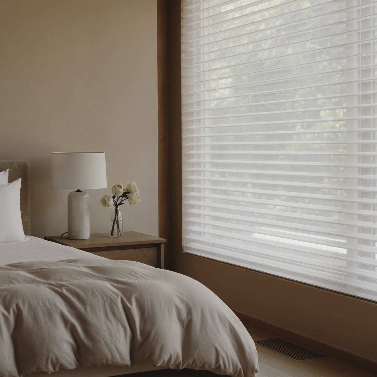White horizontal shangrila shades partially drawn on a window in a modern, minimalist bedroom with beige walls, a wooden nightstand, and a white lamp.