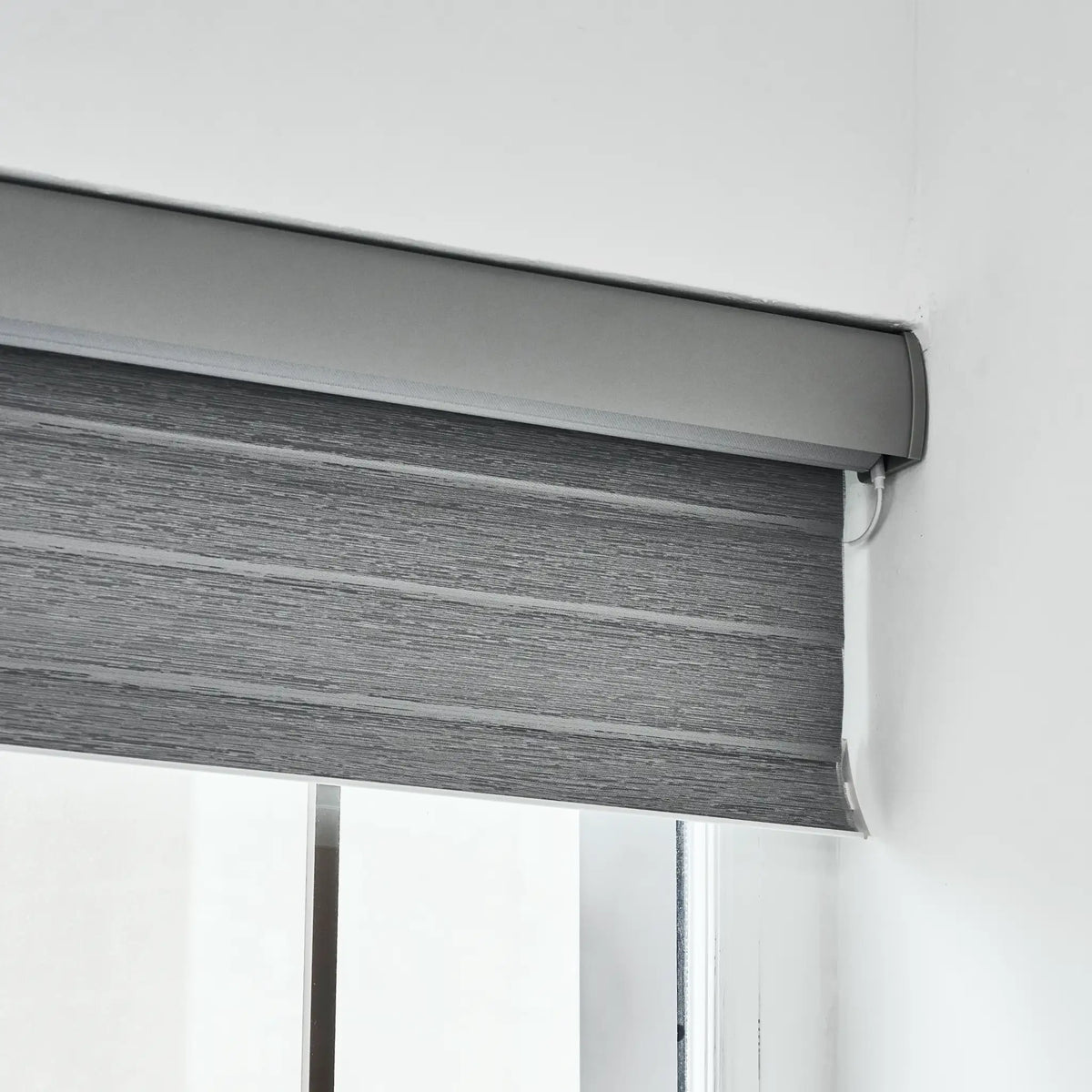 A gray roller blind with a textured horizontal slat pattern, partially rolled down and mounted on a window track against a white wall.