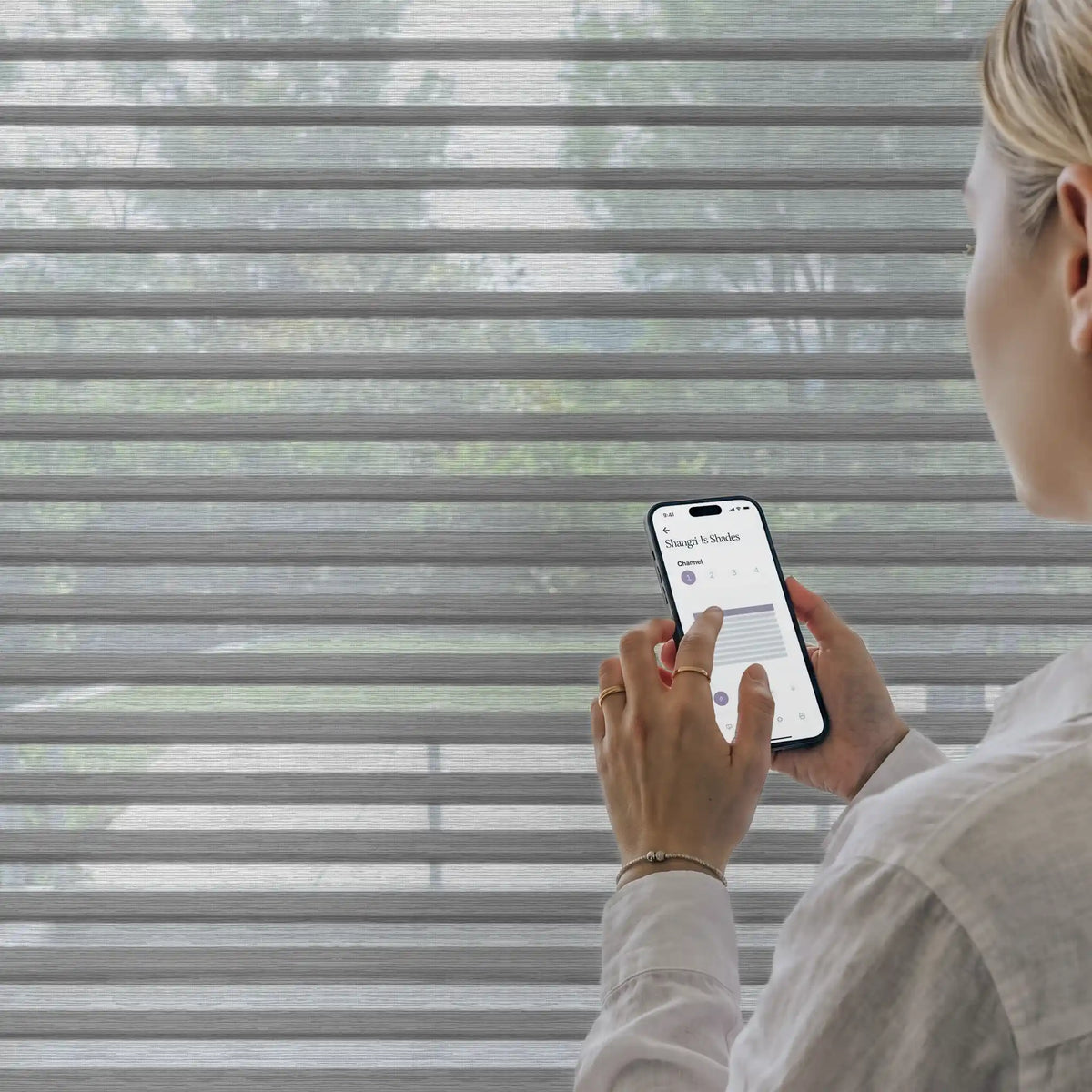 A woman in a white shirt stands in front of gray horizontal blinds, holding a smartphone that displays a smart home app interface.