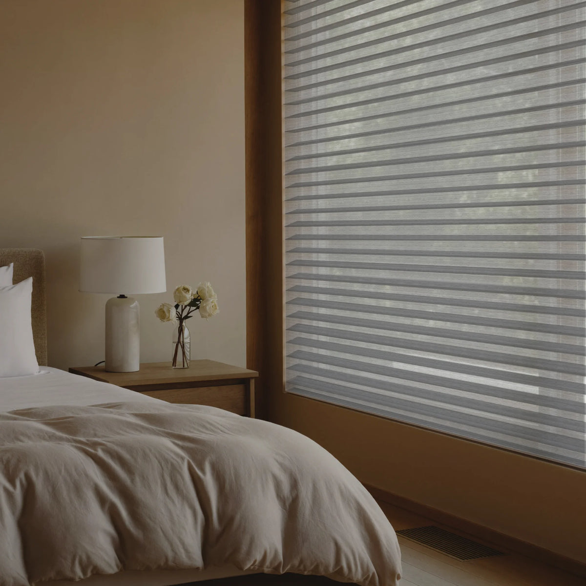 Horizontal roller blinds in a light gray color installed on a window in a modern, minimalist bedroom setting.