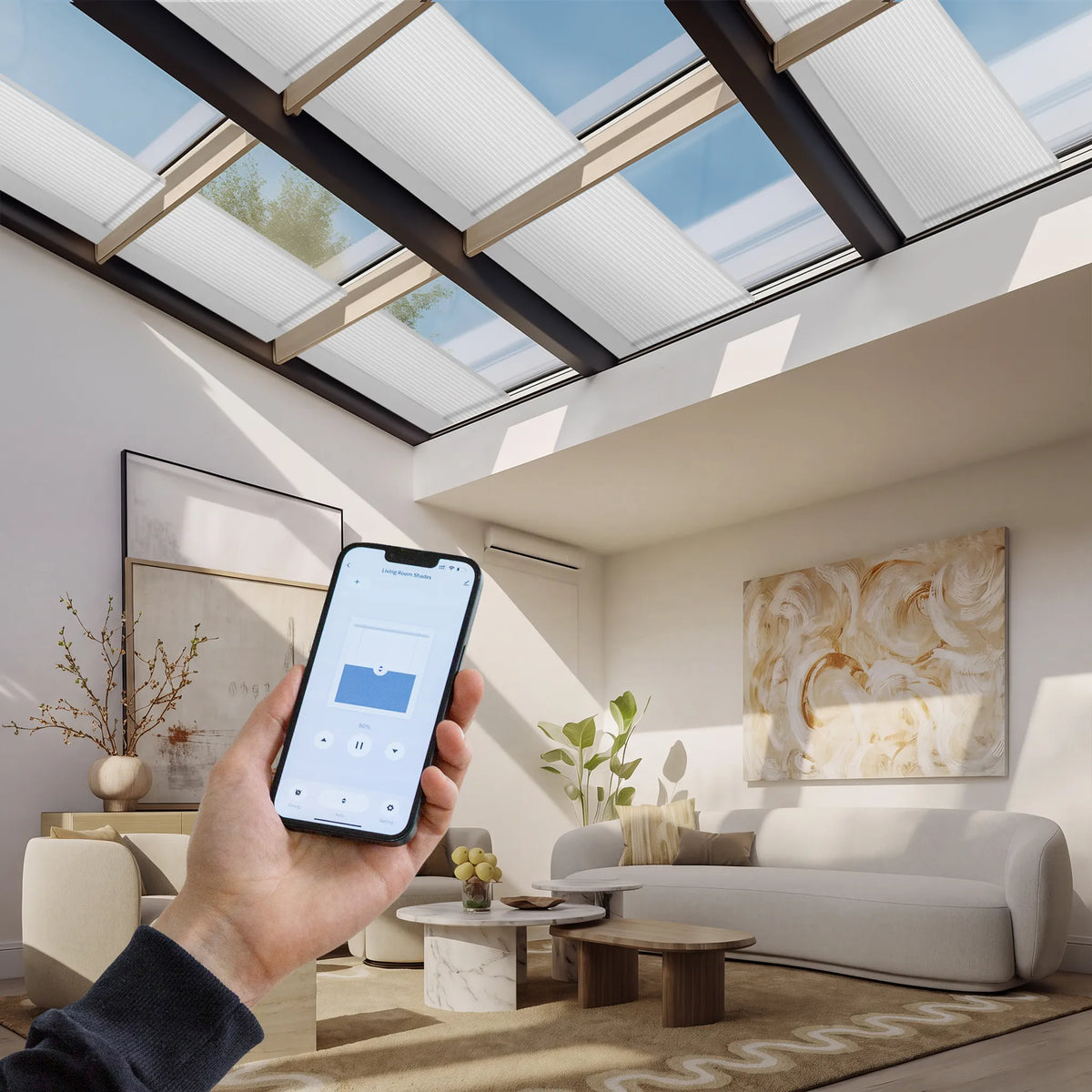 A hand holds a smartphone in a bright modern living room with a large skylight overhead. The phone screen shows a smart home control app, and the room features light-colored furniture, abstract wall art, and natural sunlight streaming through the glass roof panels.