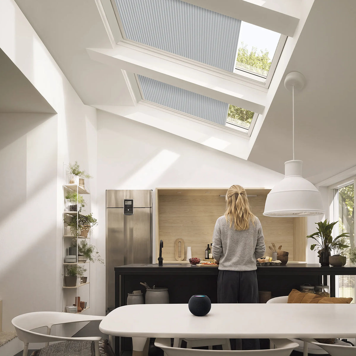 A person stands with their back to the camera in a bright, modern kitchen, with two skylights above featuring light blue-gray roller blind shades. The room has white walls, a white pendant light, and a black kitchen island.