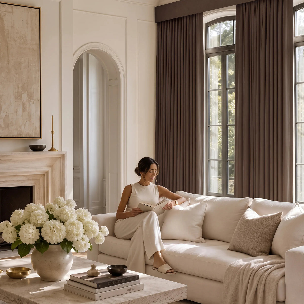 A woman sits on a cream sofa in a bright, elegant living room, reading a book. The room features dark brown floor-length curtains, a cream sofa with neutral pillows, a marble coffee table with white flowers, and a fireplace in the background.