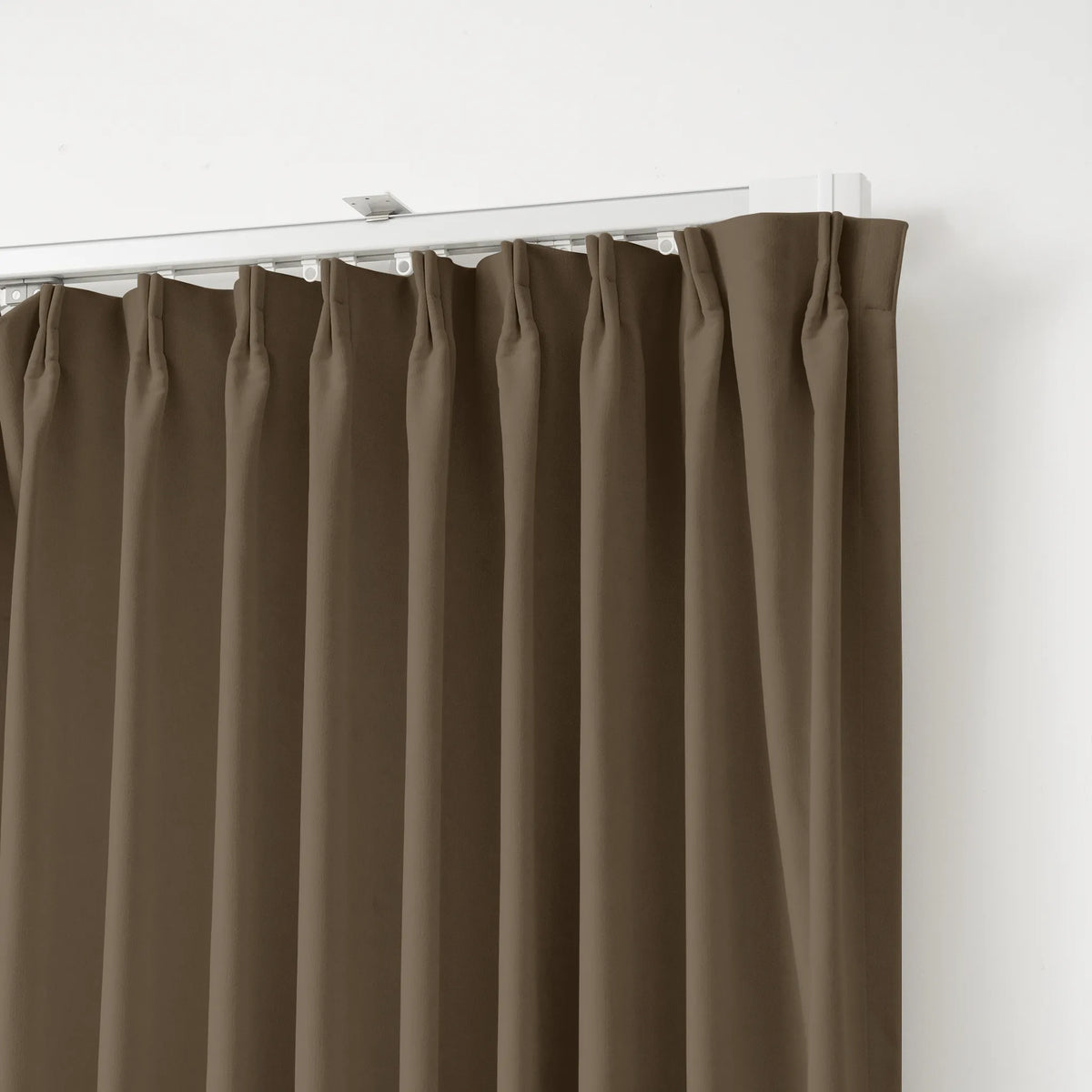 A close-up of a brown pleated curtain panel hanging from a white track against a plain white wall.