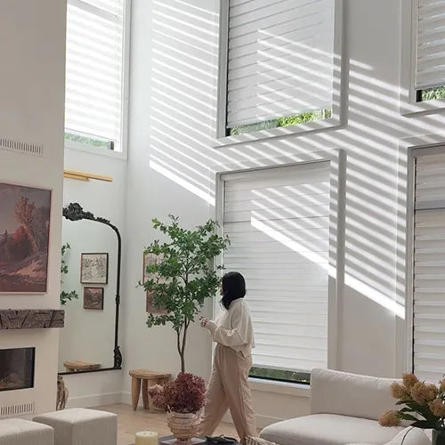 A person stands in a bright, modern living room with tall windows and white zebra blinds casting strong diagonal shadows. The room has white walls, a white sofa, a potted tree, and framed wall art.