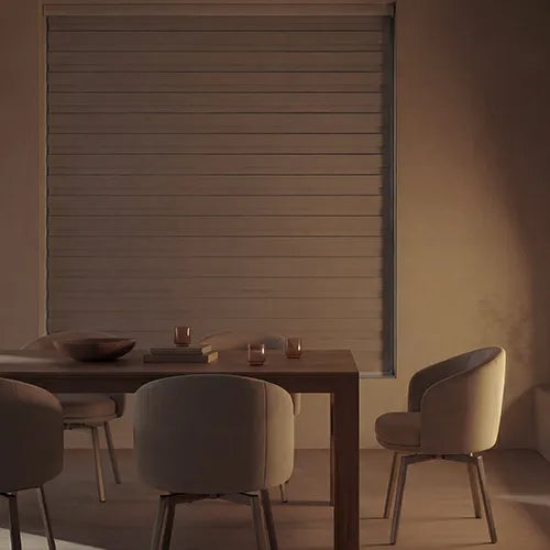A modern dining room scene with a large horizontal zebra blinds in a warm taupe-beige tone, shown behind a wooden table and upholstered chairs in a softly lit interior.