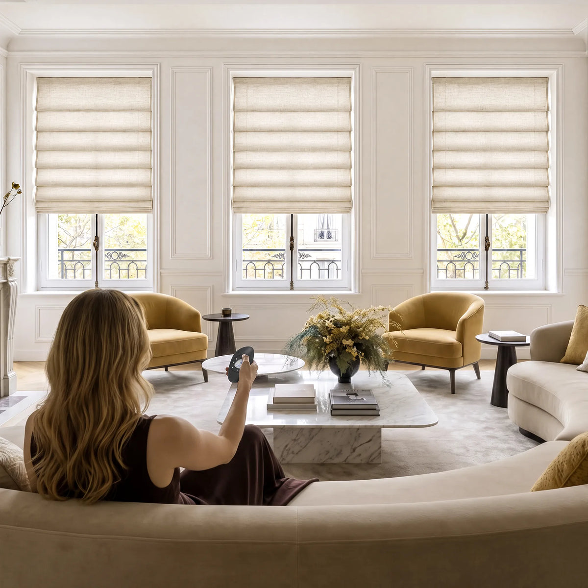 A person using a remote to adjust soft fold Roman shades across living room windows.