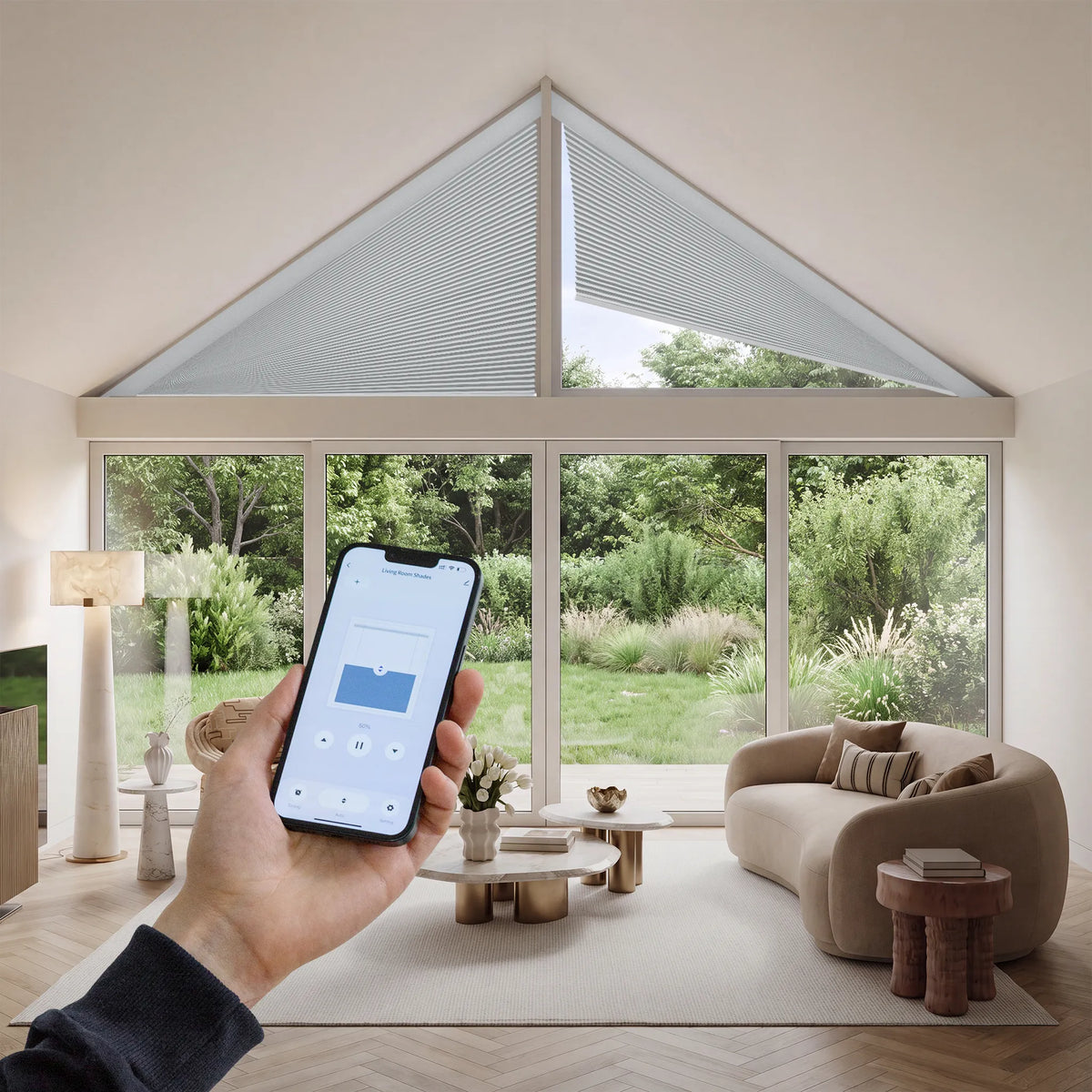 A hand holds a smartphone showing a smart home app interface in front of large triangular roof windows with white triangular cellular shades, in a bright modern living room with a curved sofa and green garden view.