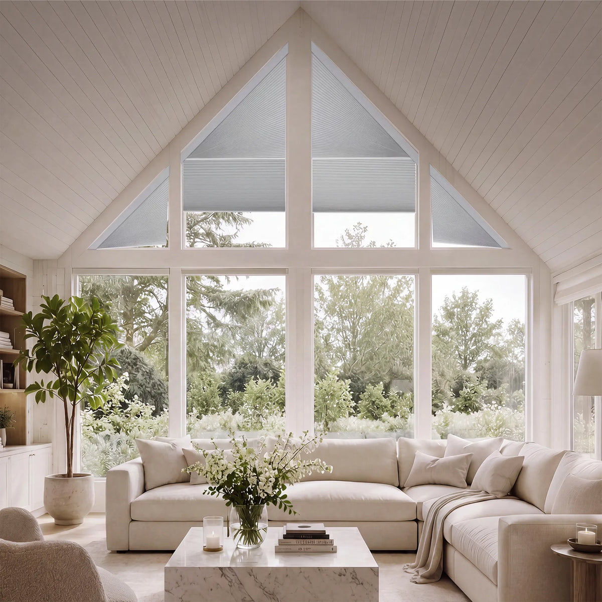 A bright, modern living room with a large cream sectional sofa, a marble coffee table, and a tall potted plant in front of a large multi-pane triangular window looking out to green trees.